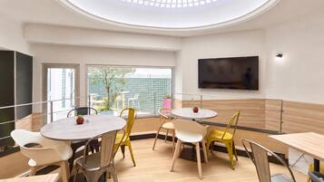 A modern dining area with round tables and chairs, a flat-screen TV mounted on the wall, and a view of the outdoors through large windows.