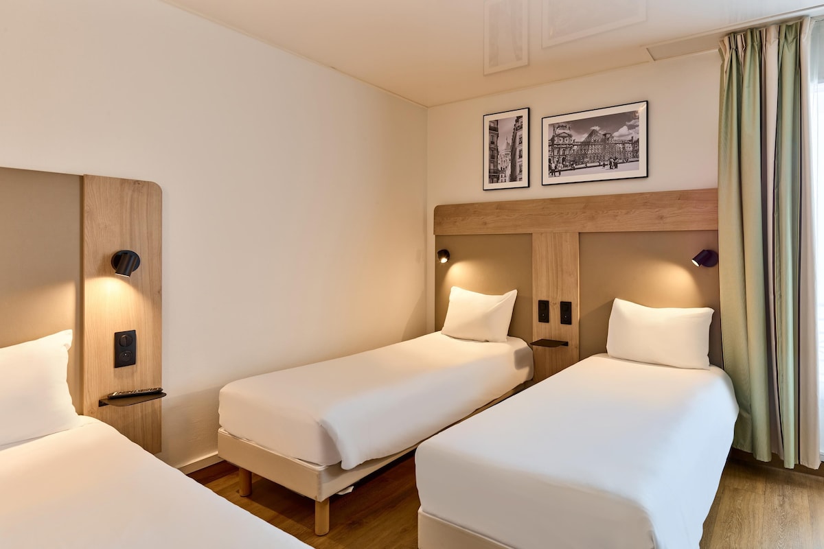 A hotel room with two single beds, a wooden headboard, a wall-mounted light, and a window with curtains.