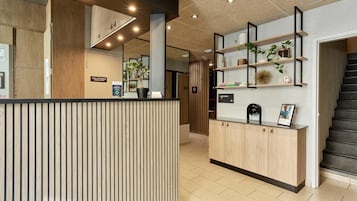 Un espace d’accueil avec un comptoir en bois, une signalétique indiquant l’emplacement d’une salle technique locale, et un escalier sur la droite.