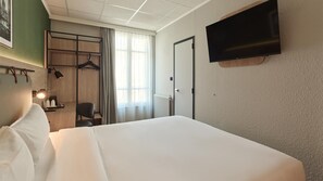 A hotel room with a bed, a desk with a chair, a TV mounted on the wall, and a window with curtains. - Campanile PRIME - Paris Ouest Boulogne (Boulogne-Billancourt)