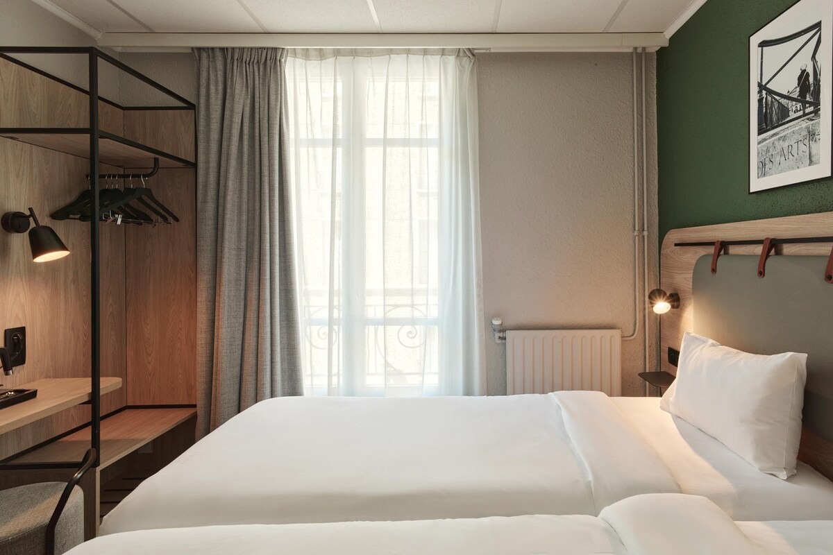 Une chambre d’hôtel avec un lit, un bureau, une chaise, un radiateur et une fenêtre avec des rideaux.