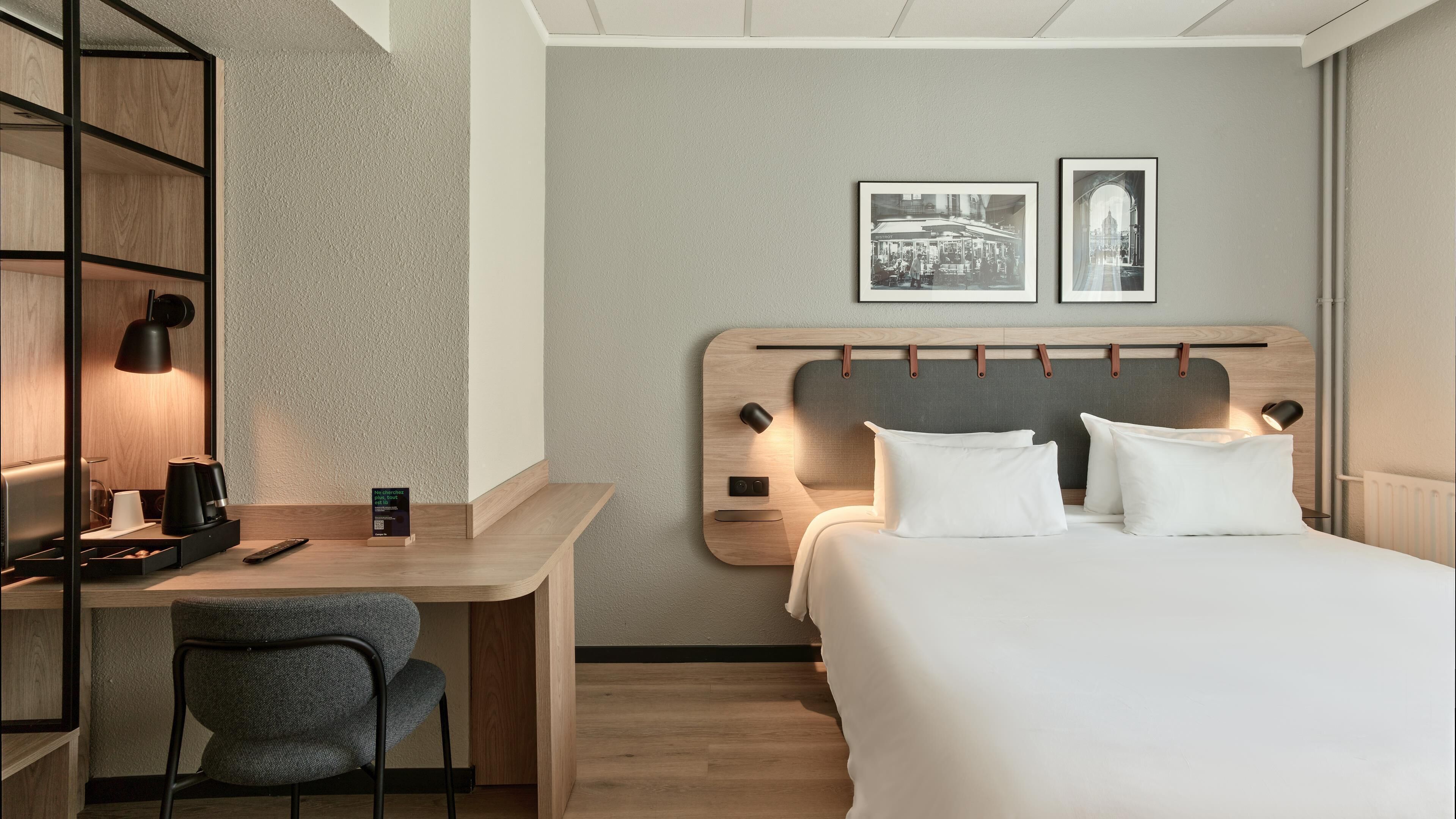 A hotel room with a bed, a desk, a chair, a coffee maker, and two framed pictures on the wall.