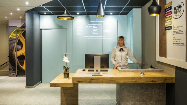 A reception desk with a modern design, a potted orchid, and a digital display in the background. - ibis Clichy Centre Mairie (Clichy)