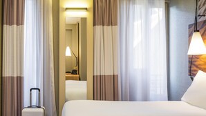 A hotel room with a bed, curtains, a lamp, and a suitcase. - ibis Clichy Centre Mairie (Clichy)