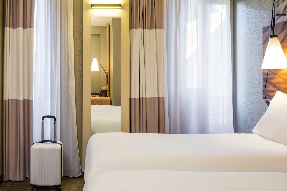 A hotel room with a bed, curtains, a lamp, and a suitcase.