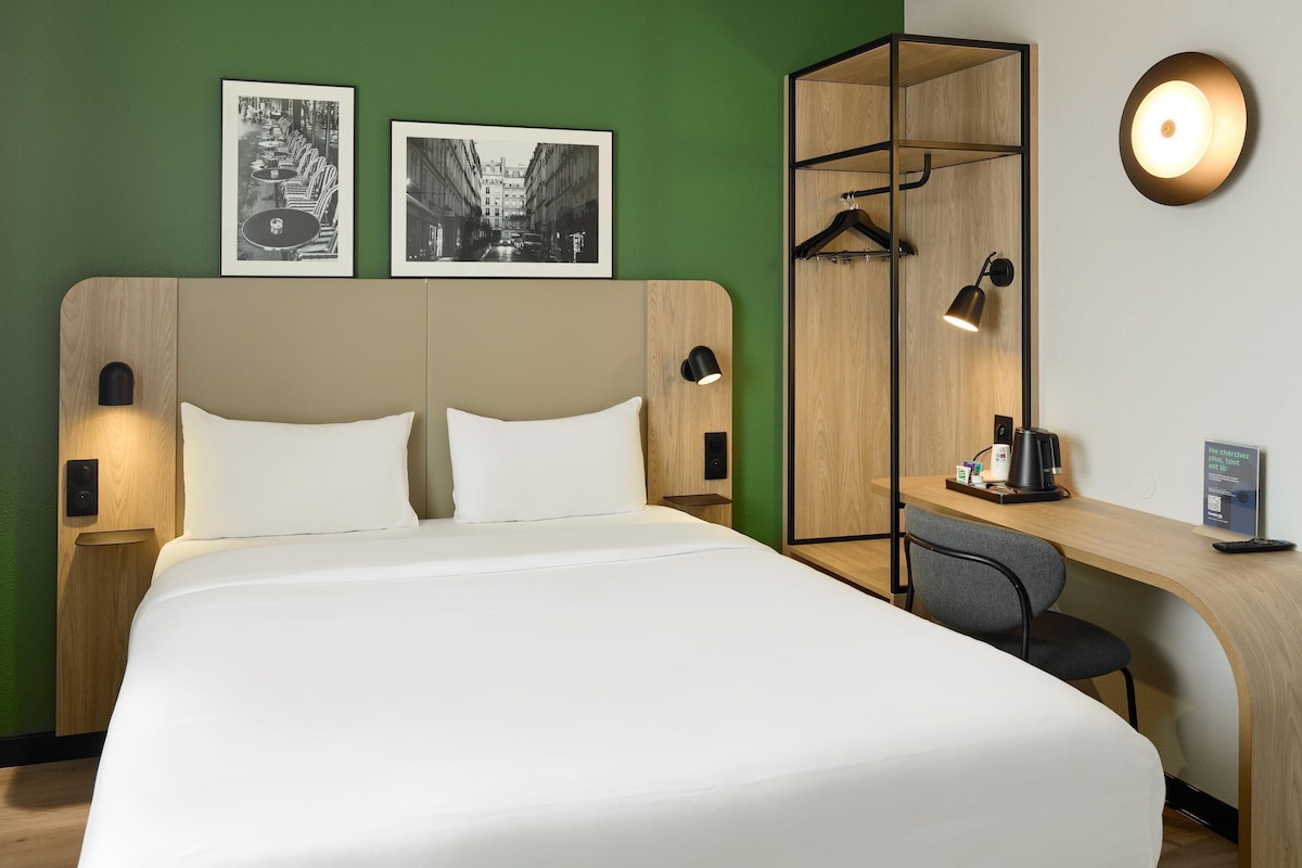 Habitación de hotel con cama, escritorio, silla, armario y dos cuadros en la pared.