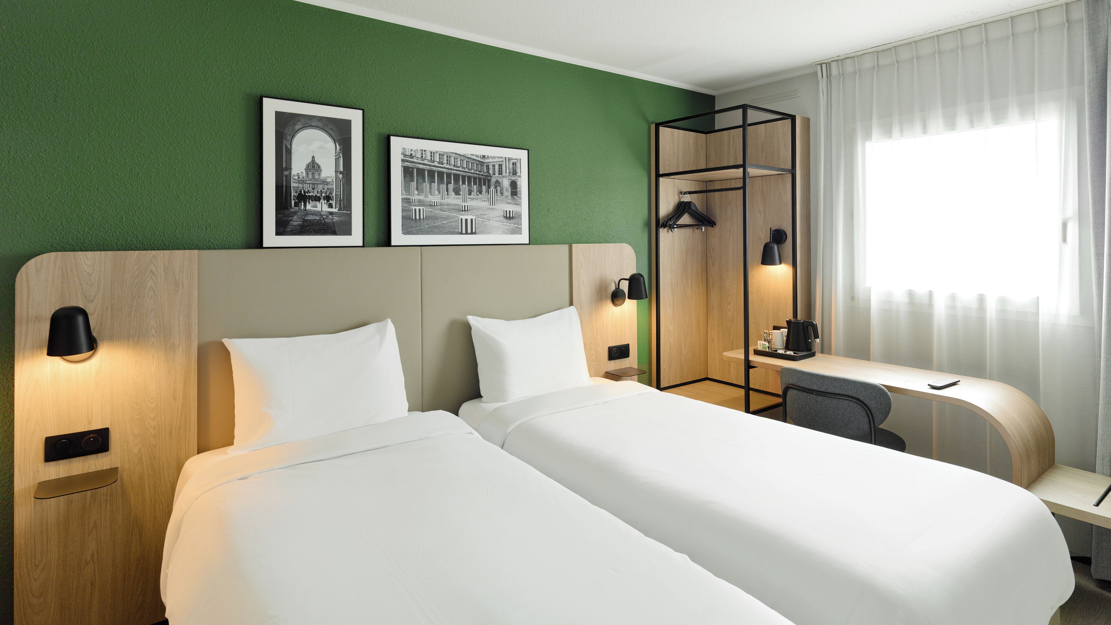 A hotel room with two beds, a desk, a chair, a coat rack, and two framed pictures on the wall.