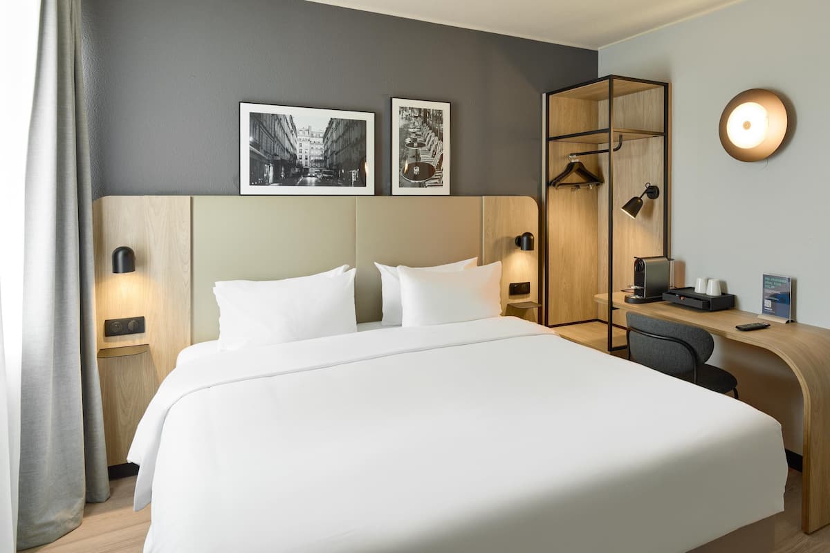 A hotel room with a large bed, a desk with a computer, a mirror, and two framed pictures on the wall.