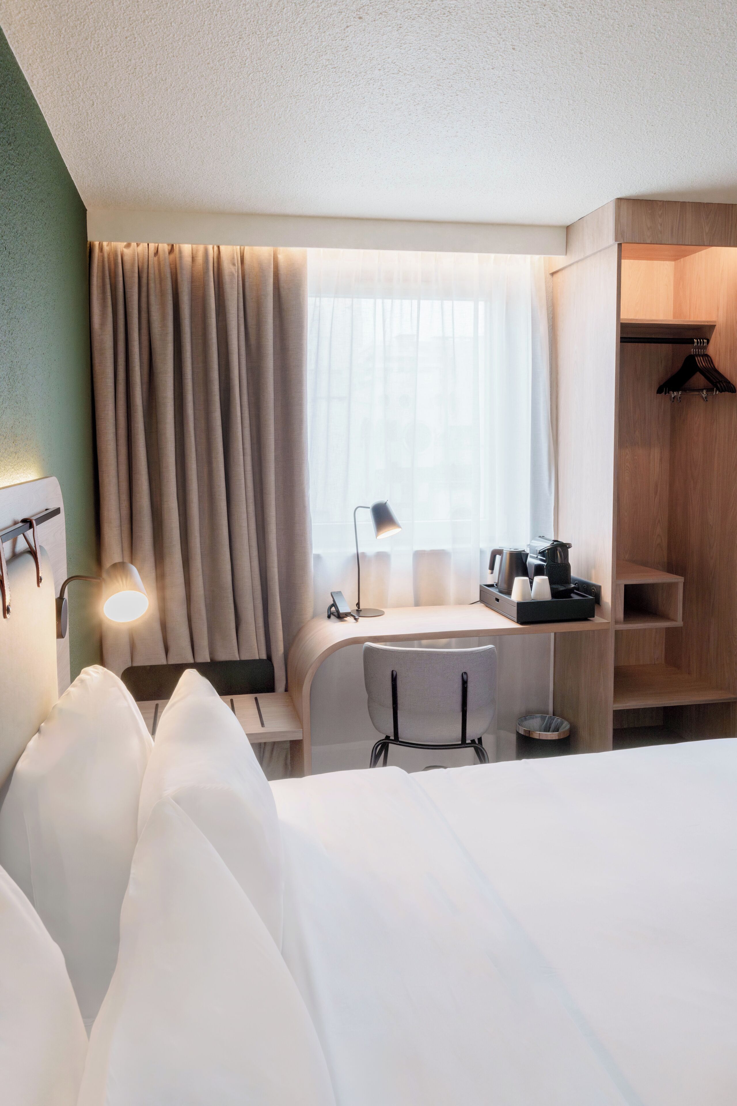 Habitación de hotel con cama, escritorio, silla, lámpara y un pequeño rincón de cocina.