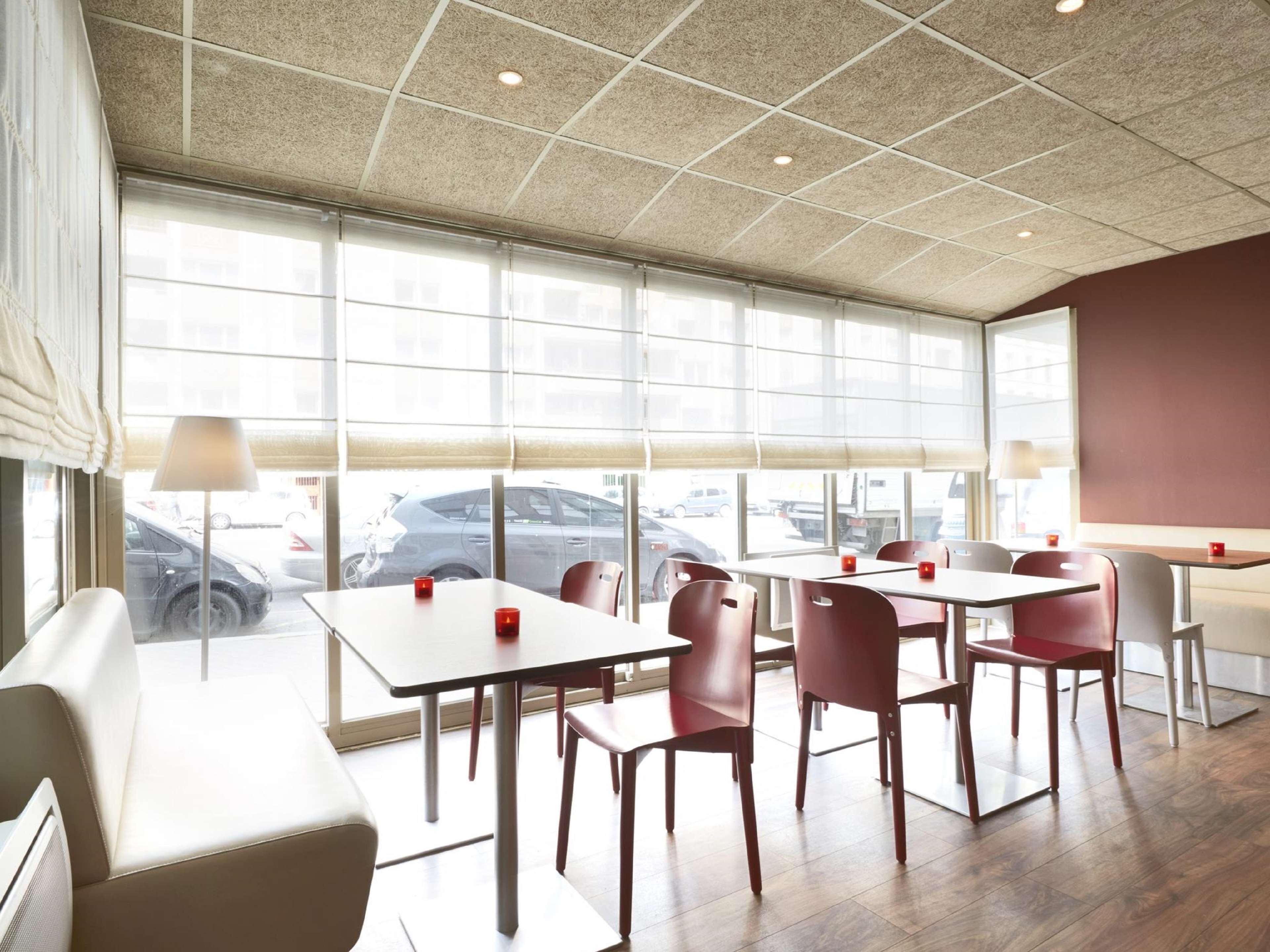 A modern dining area with large windows, a high ceiling, and a view of parked cars outside.