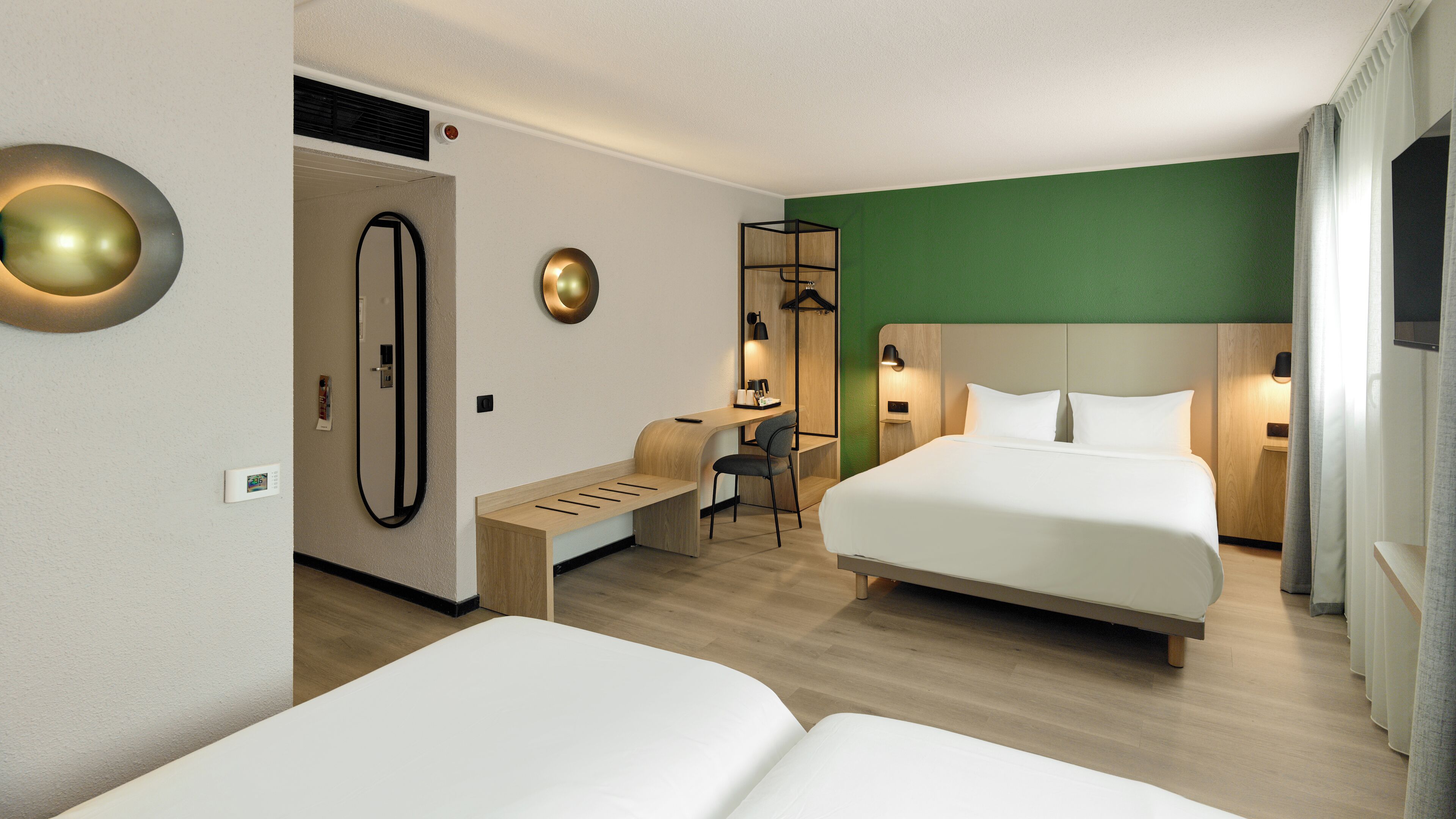A hotel room with two beds, a desk, a chair, a mirror, and a green accent wall.