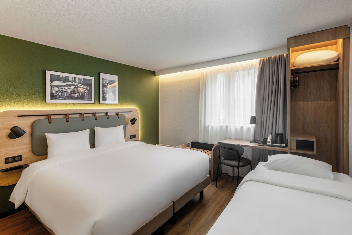 Habitación de hotel con una cama grande, un escritorio y una silla. Hay dos cuadros en la pared, una ventana con cortinas y un mueble de madera con espejo.