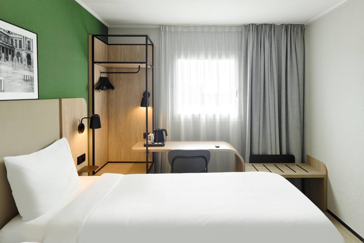 A hotel room with a bed, a desk, a chair, a lamp, and a window with curtains.