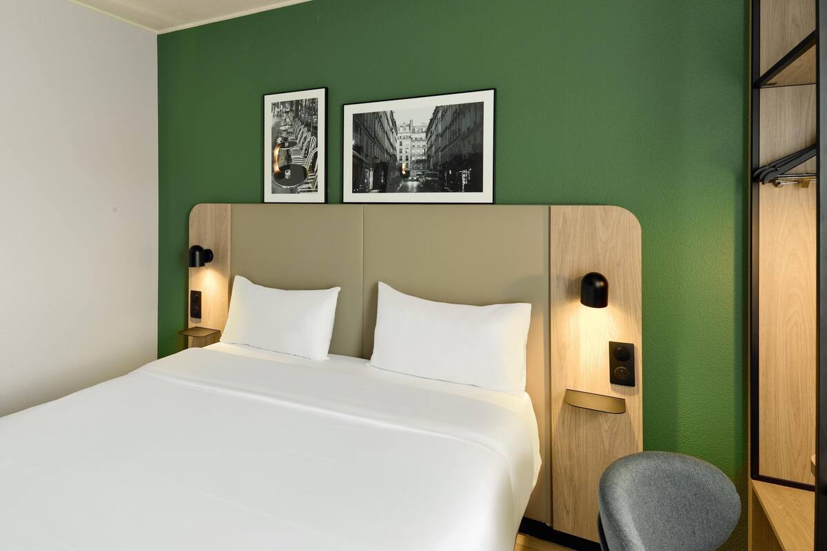 A hotel room with a bed, two wall-mounted picture frames, a round wall light, and a wooden headboard.