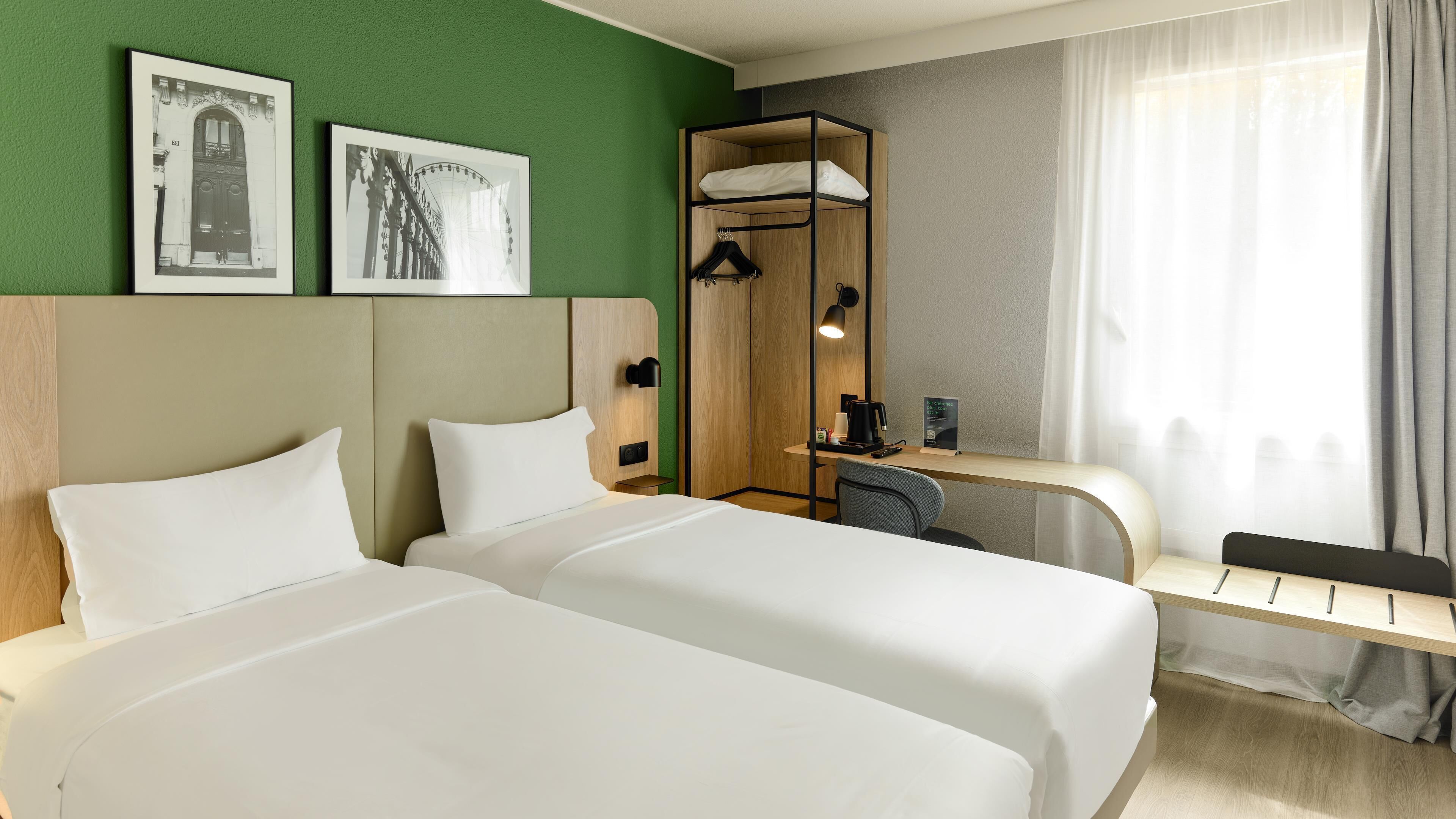 A hotel room with two beds, a desk, a chair, a shower area, and two framed pictures on the wall.