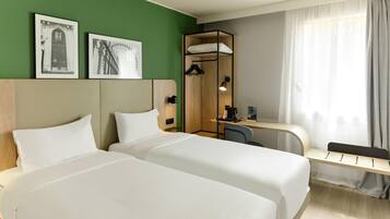 A hotel room with two beds, a desk, a chair, a shower area, and two framed pictures on the wall.