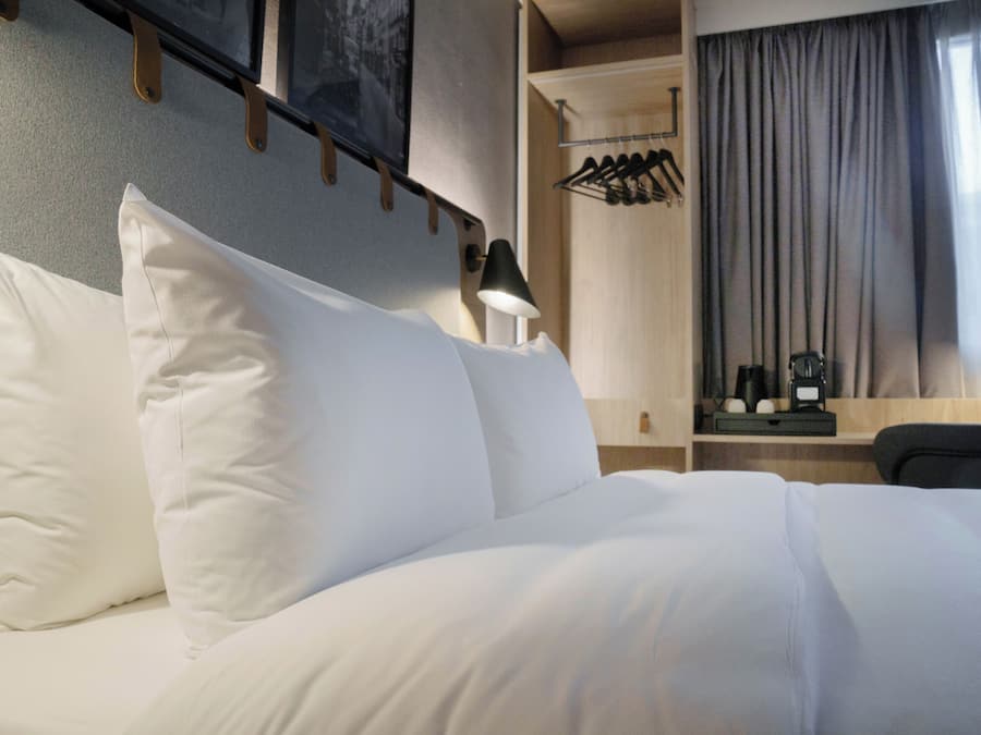 Una cama bien hecha con sábanas blancas, una lámpara de pared y un escritorio de madera con un espejo y un pequeño reloj.