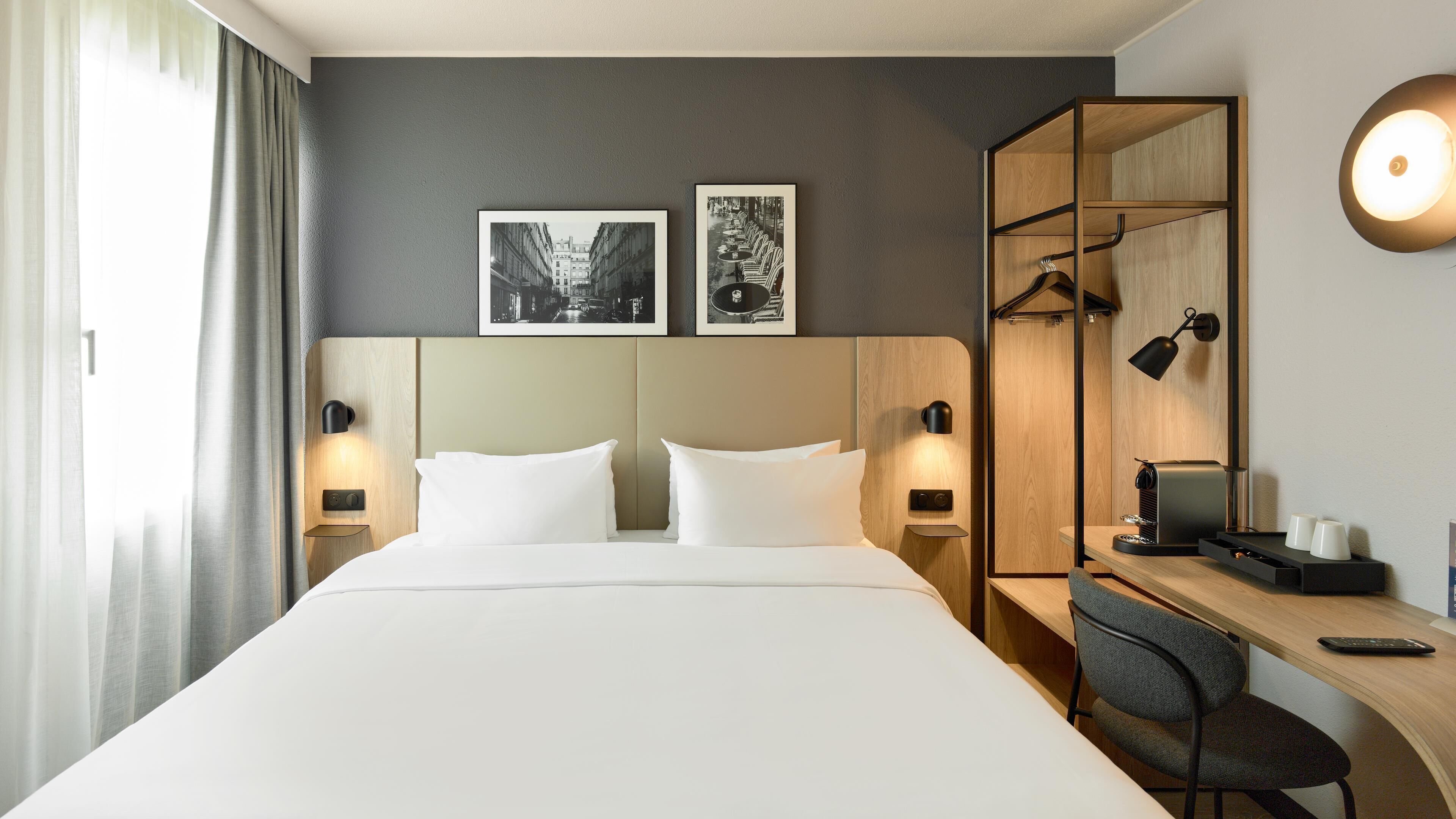 A hotel room with a large bed, two bedside tables with lamps, a desk with a chair, and two framed pictures on the wall.