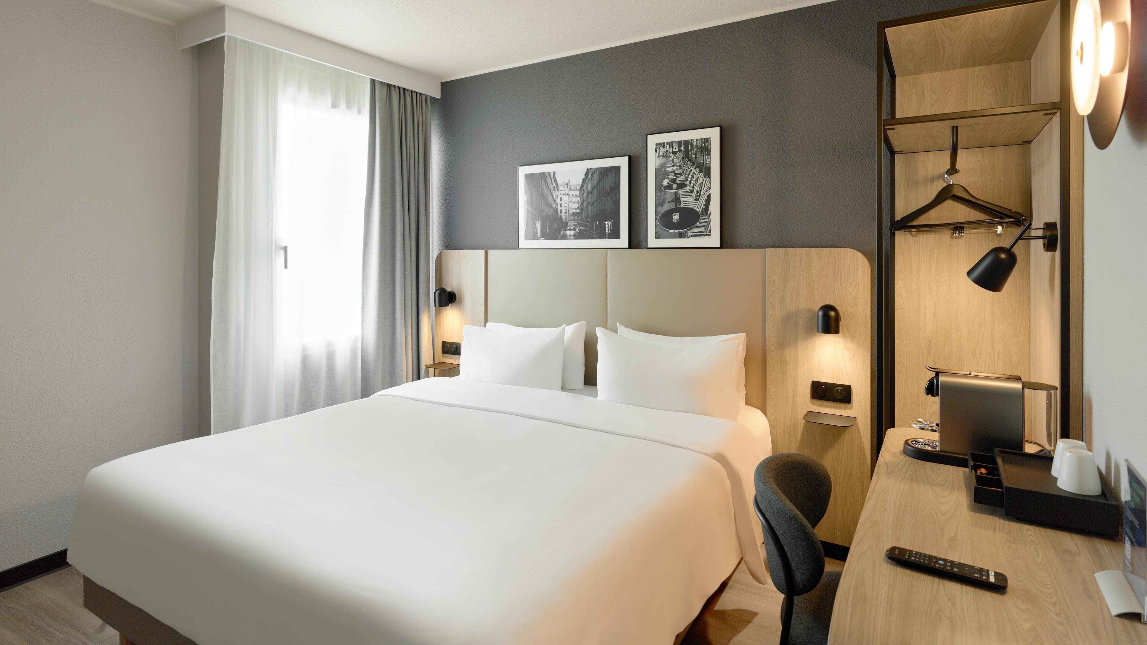A hotel room with a large bed, a desk with a coffee machine, a chair, a desk lamp, and two framed pictures on the wall.