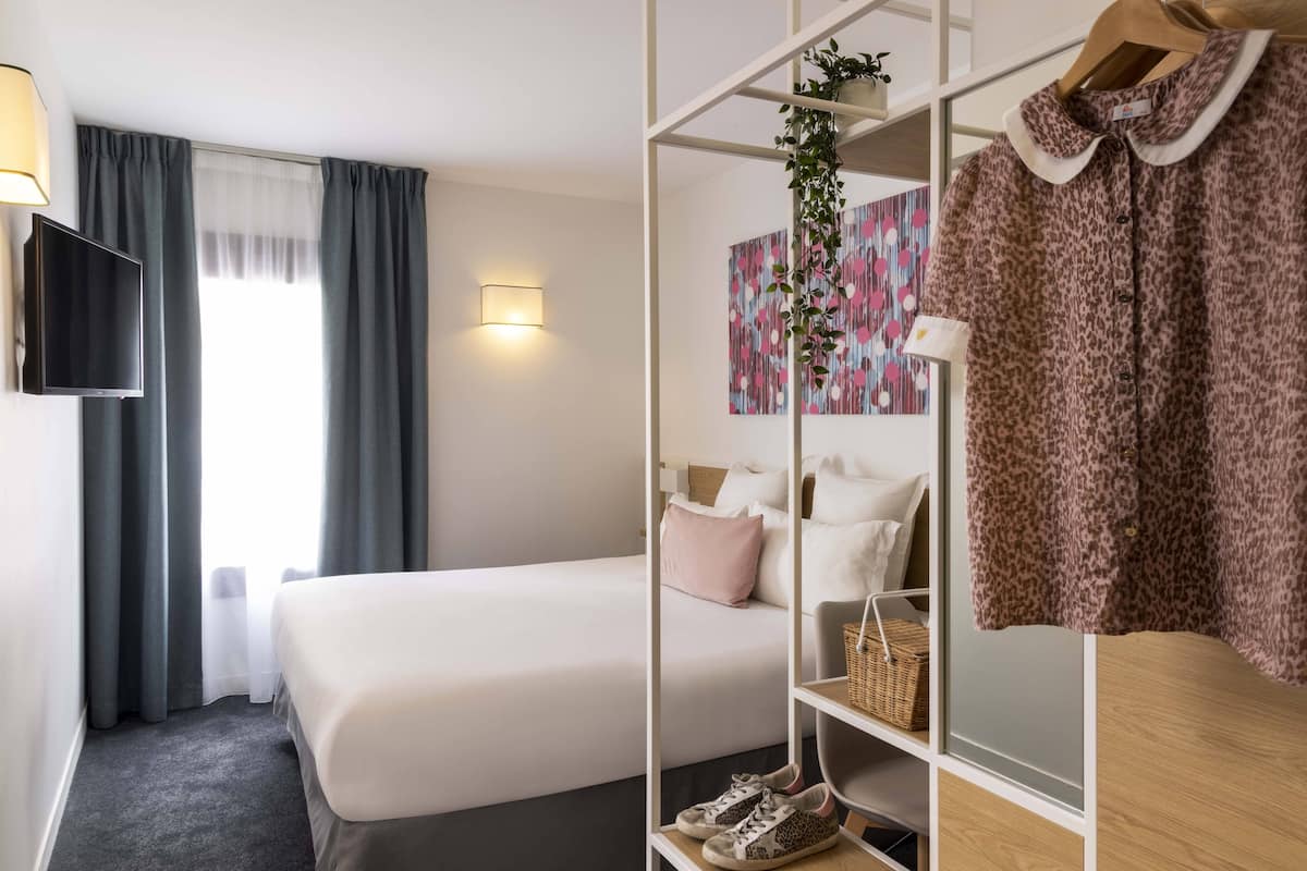 Habitación de hotel con cama, televisión, armario y estante con zapatos y una cesta.