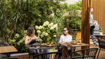 Un café en plein air, avec des tables et des chaises, entouré d’une végétation luxuriante et de fleurs blanches.