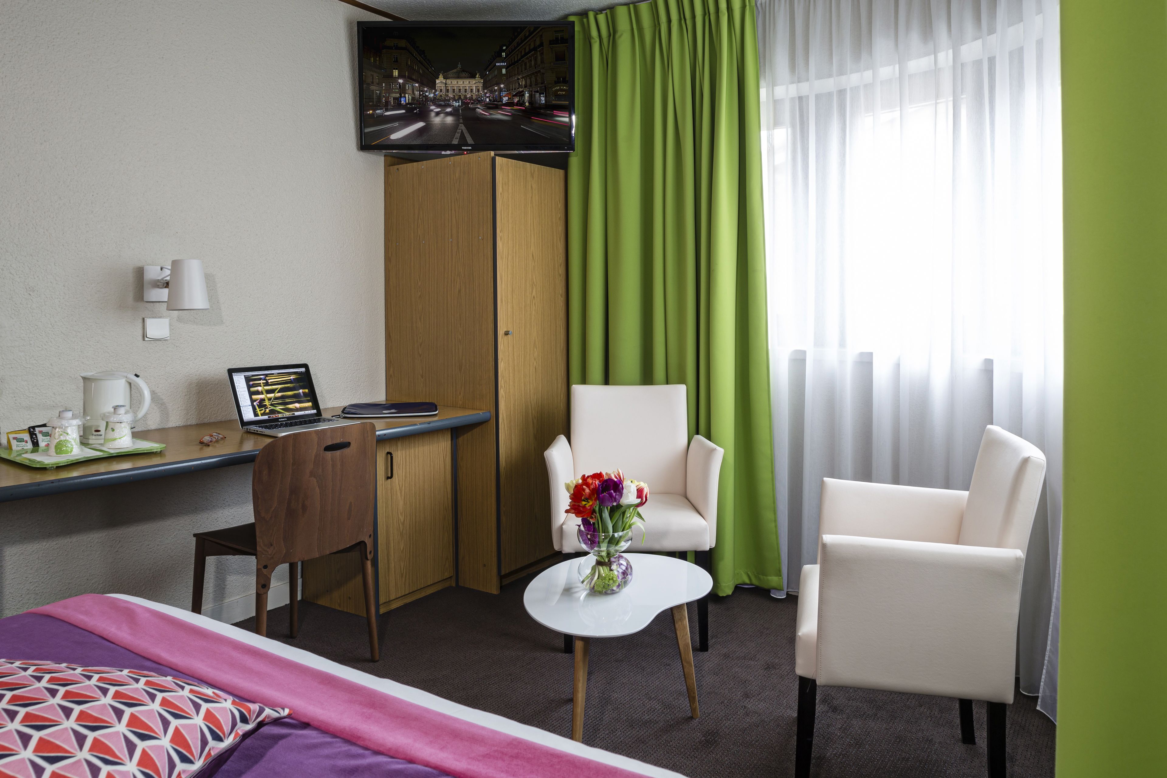 Une chambre d’hôtel comprenant un lit, deux chaises, un bureau avec un ordinateur portable, une machine à café et un vase avec des fleurs.