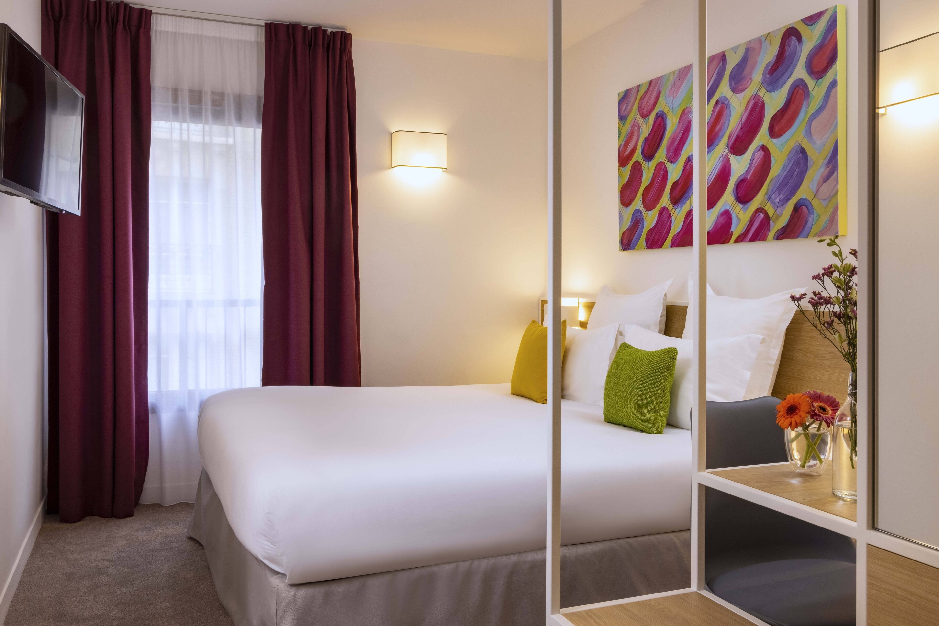 A hotel room with a bed, a TV, a painting, and a vase with flowers.
