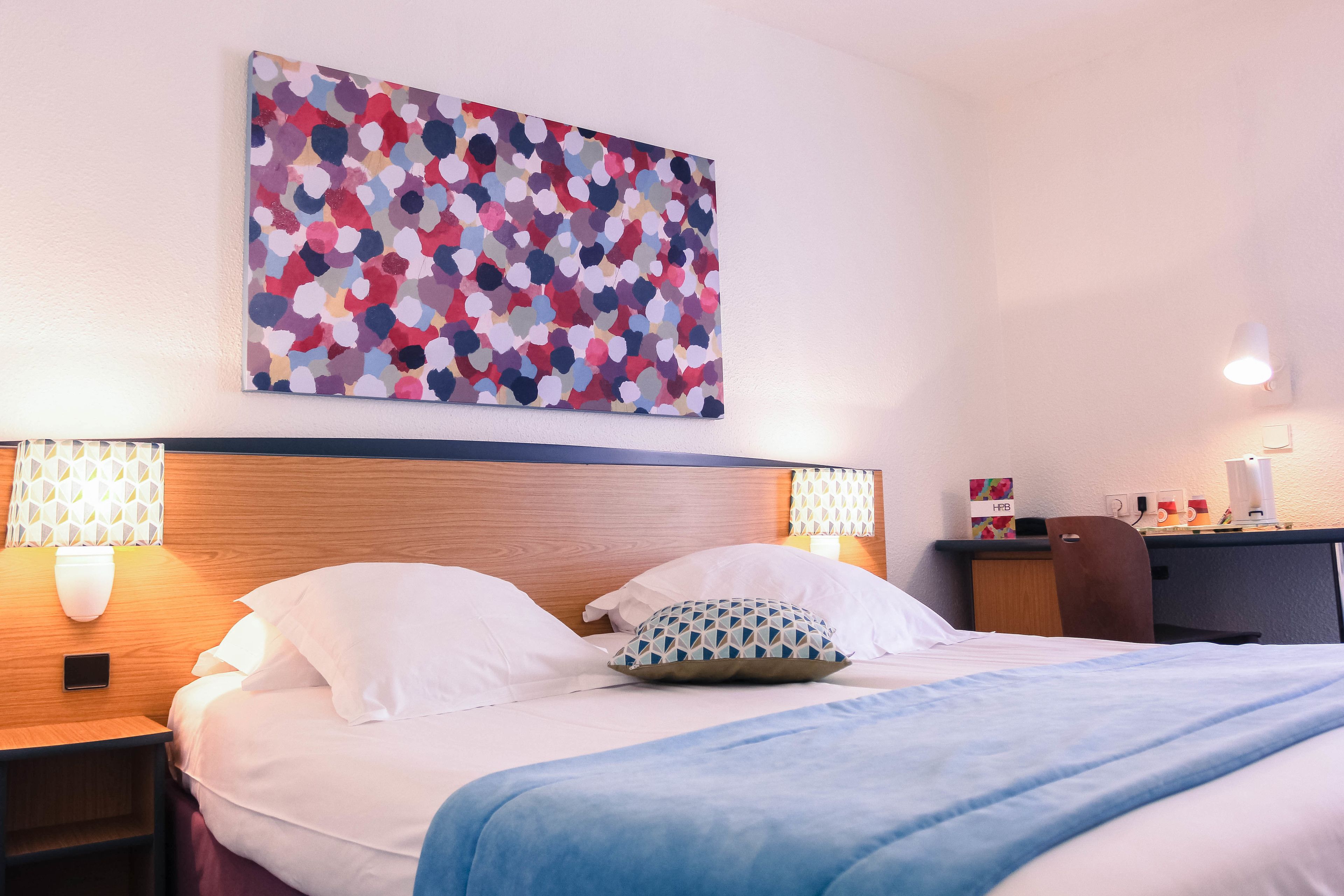 Una cama bien hecha con ropa de cama blanca y azul, un cuadro abstracto colorido encima, y un escritorio con una lámpara y una silla a la derecha.