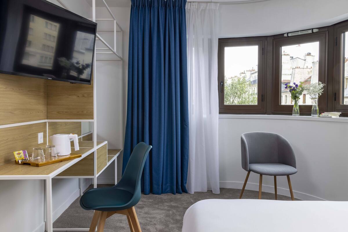 Habitación de hotel con cama, escritorio con cafetera, silla, televisor montado en la pared y ventana con cortinas.