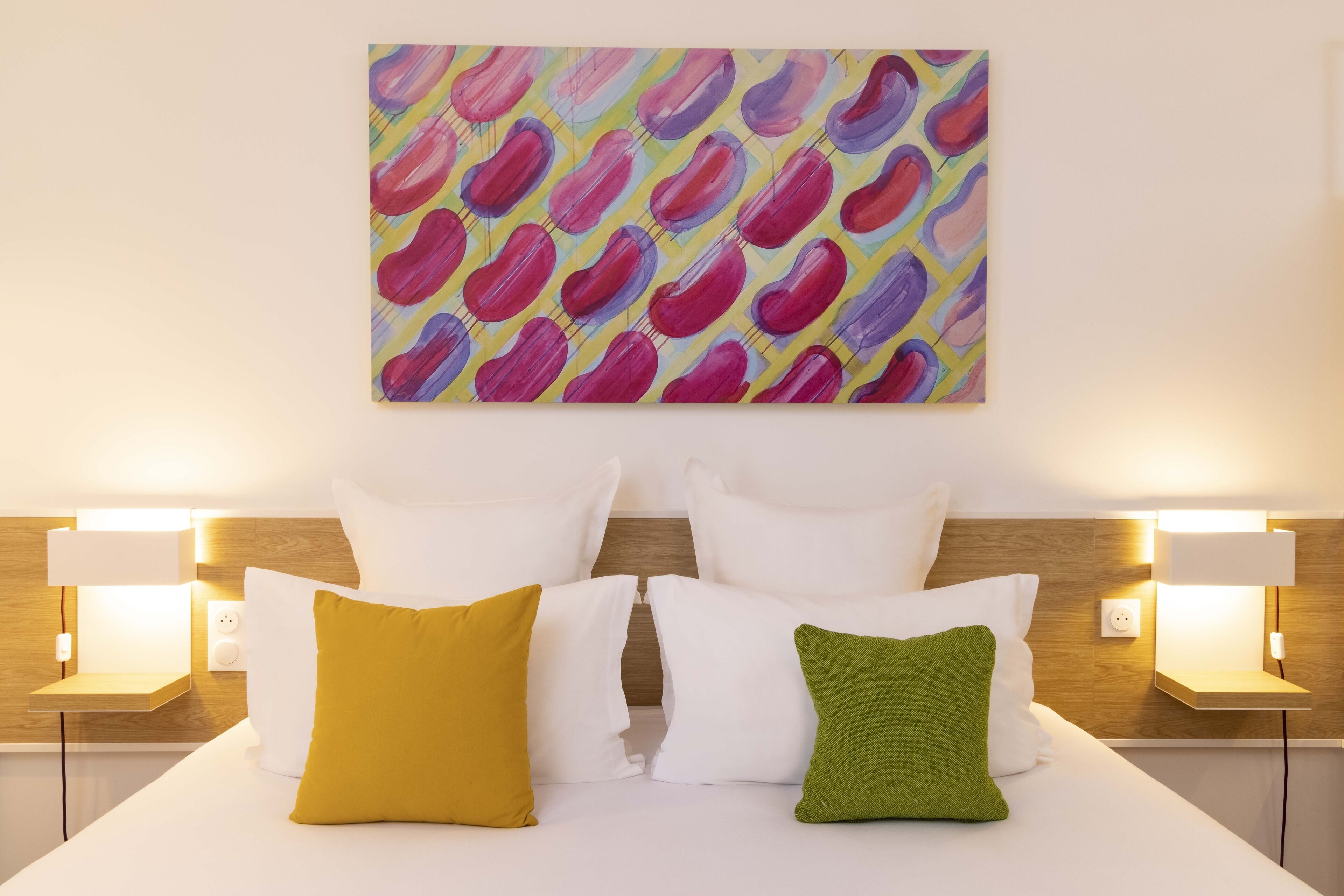 Une chambre d’hôtel avec un lit, des oreillers blancs et verts, et un tableau abstrait aux couleurs vives accroché au-dessus du lit.
