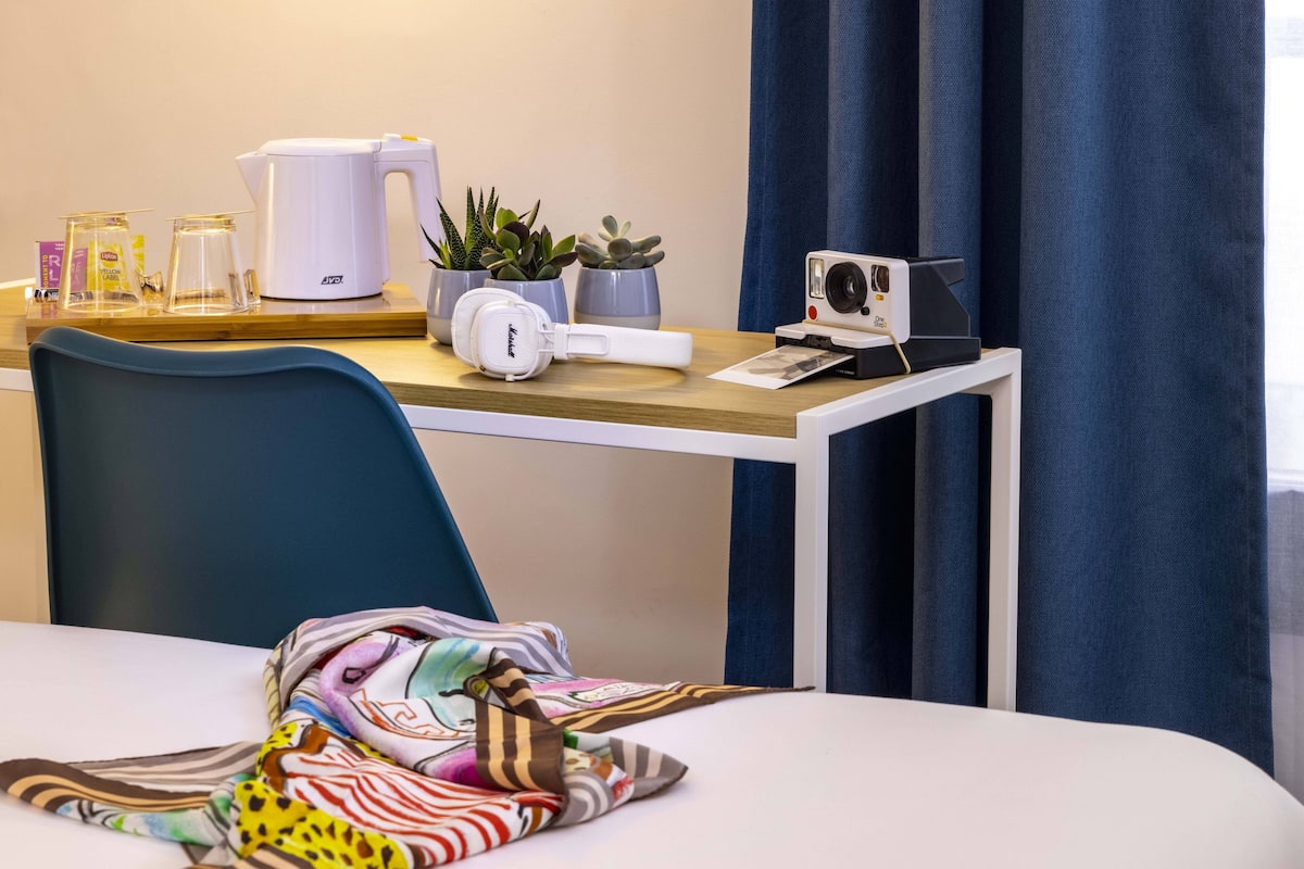 Habitación de hotel con un escritorio de madera, una tetera blanca, una cámara y plantas en macetas.