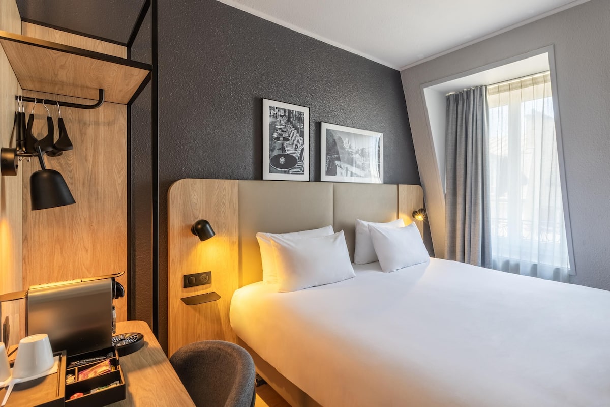 A hotel room with a bed, a desk with a computer, a coffee machine, and two framed pictures on the wall.