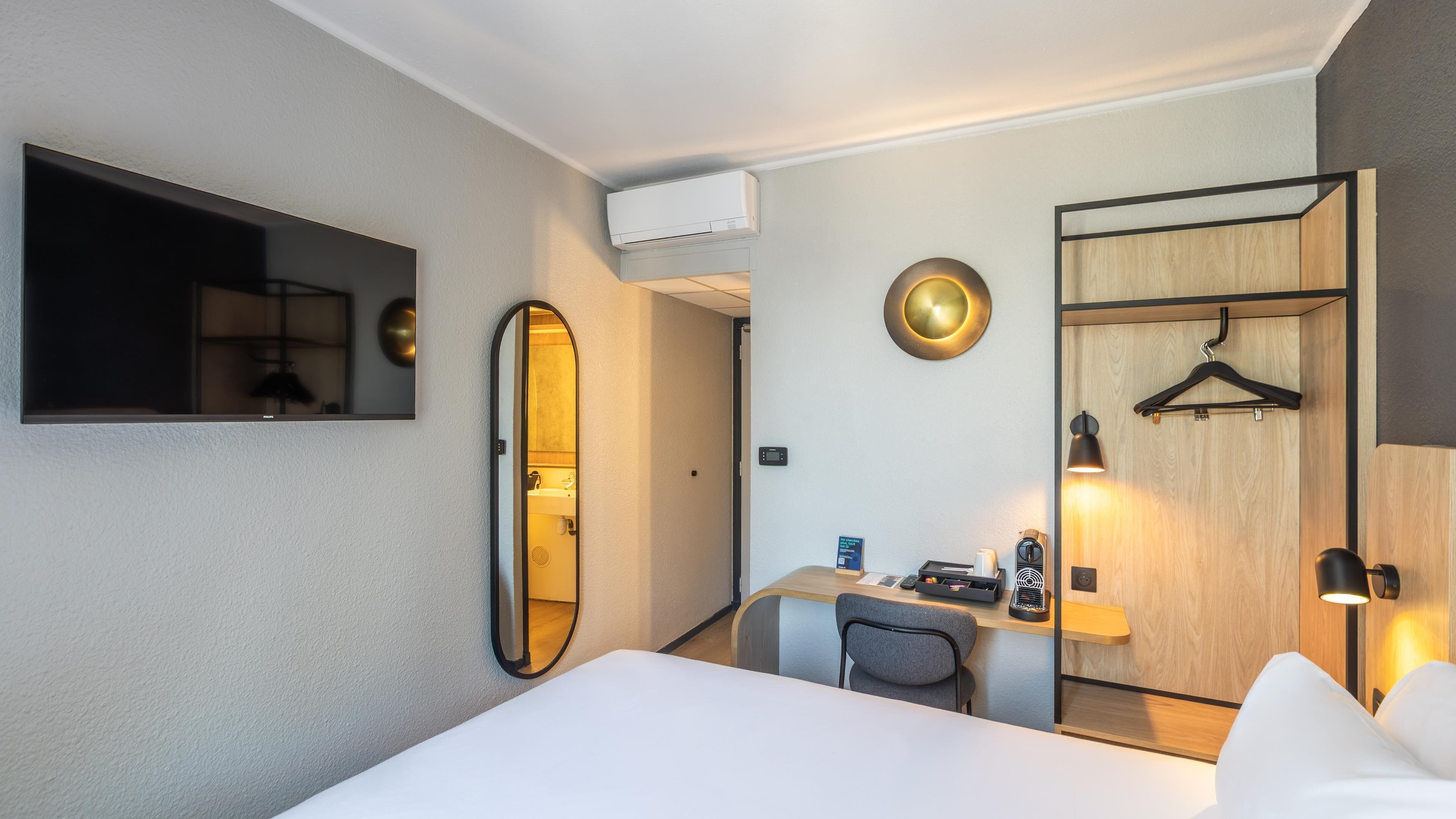 A hotel room with a bed, a desk, a chair, a mirror, a coat rack, and a wall-mounted air conditioner.