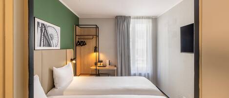 Habitación de hotel con dos camas, un escritorio con lámpara, perchero, un cuadro enmarco y televisión.