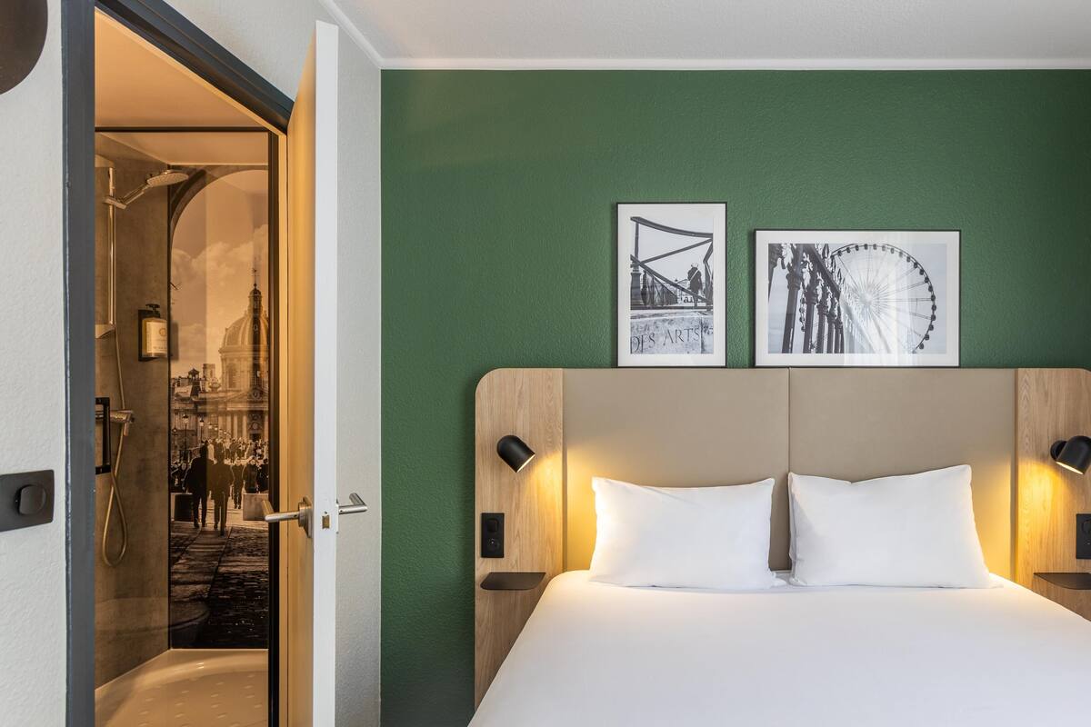 A hotel room with a bed, a green wall, two framed pictures, and a door leading to a bathroom.