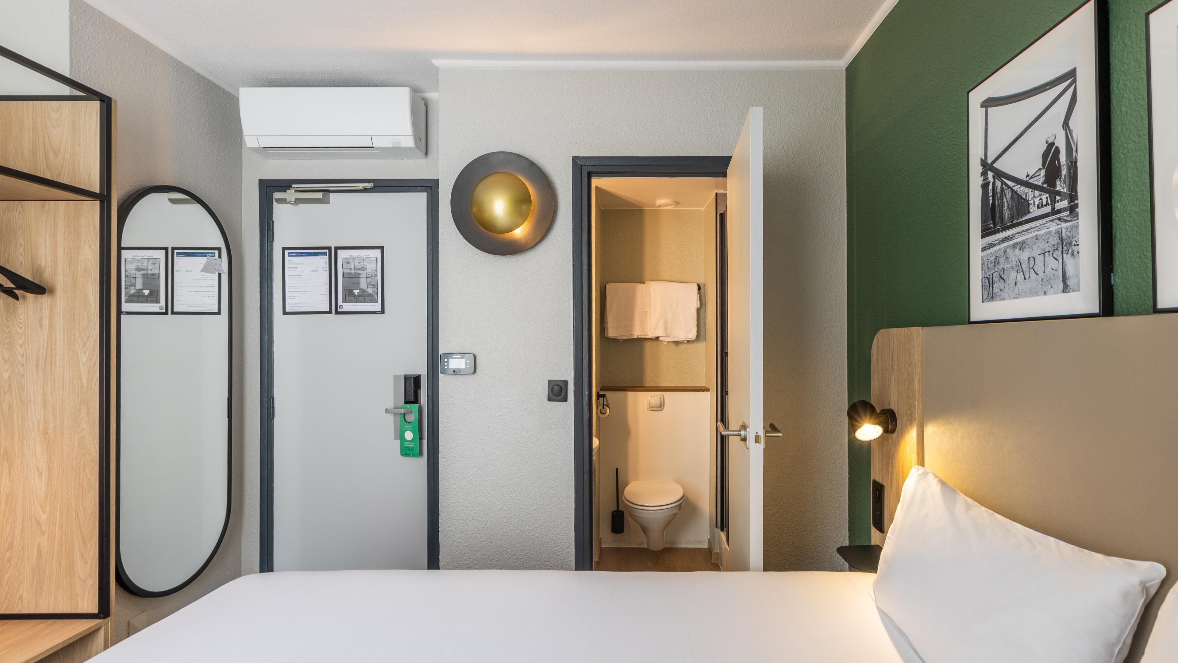 A hotel room with a bed, a door with a mirror, a wall-mounted air conditioner, a framed picture, and a toilet visible through an open door.