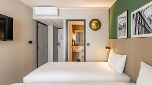 Une chambre d’hôtel comprenant un lit, une télévision, un appareil de climatisation, une salle de bain avec un lavabo et un miroir, ainsi que des tableaux encadrés accrochés au mur.
