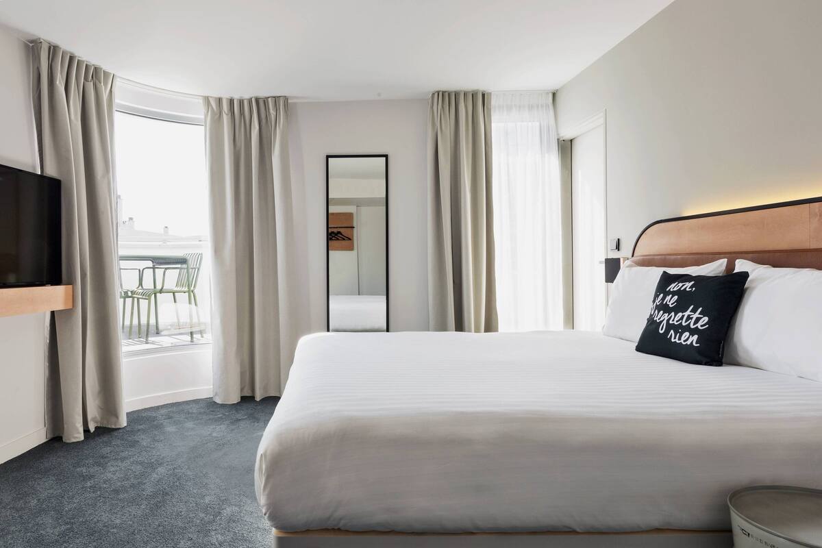 Habitación de hotel con cama, televisión de pantalla plana, espejo y balcón con sillas.