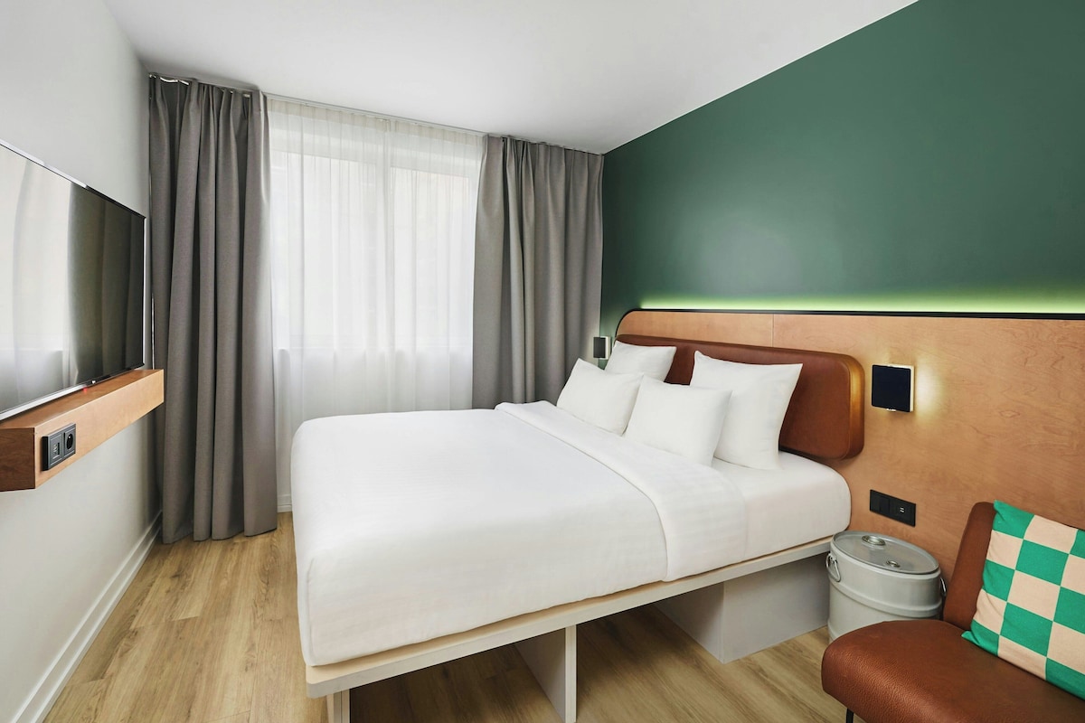 A modern hotel room with a bed, a TV, a small table, and a trash bin.