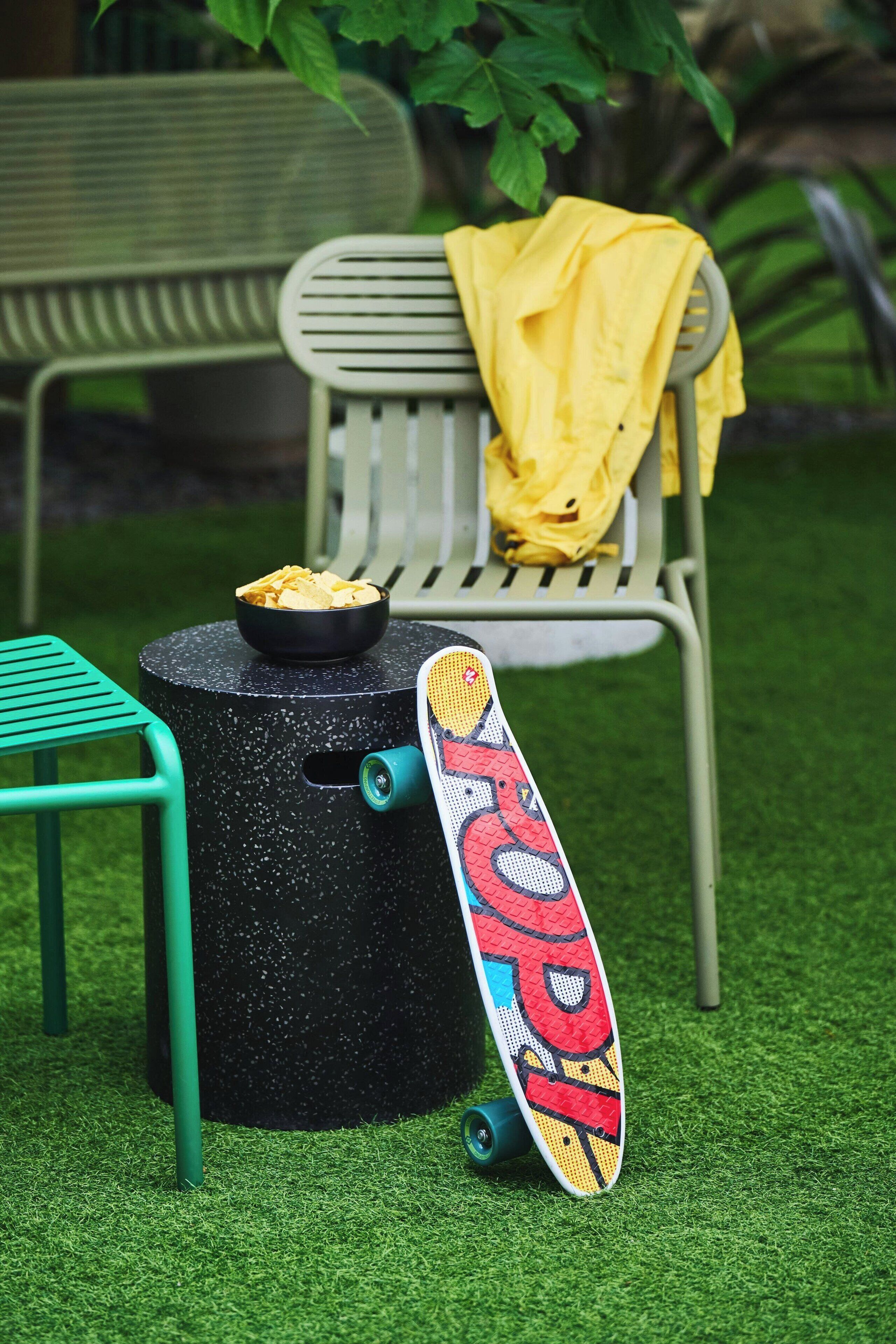 Un skateboard aux motifs colorés, un bol de chips et une chaise recouverte d’un tissu jaune, le tout posé sur une surface d’herbe artificielle verte.