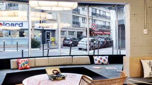 A modern interior with a round coffee table, wicker chairs, and a large window offering a view of the street and buildings outside. - Moxy Paris Bastille France (Paris)