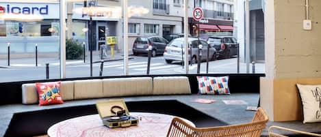 A modern interior with a round coffee table, wicker chairs, and a large window offering a view of the street and buildings outside.