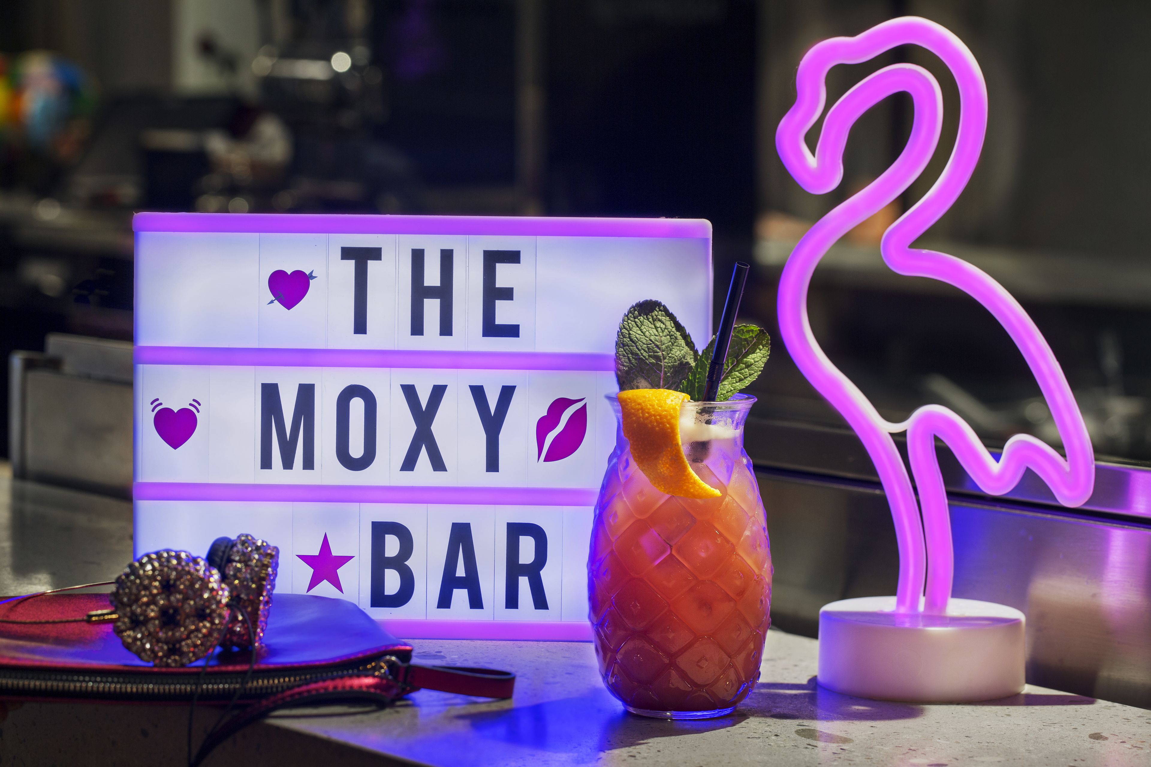 Ein Bar-Schild mit der Aufschrift "THE MOXY BAR" und einer Neon-Flamingo-Dekoration.