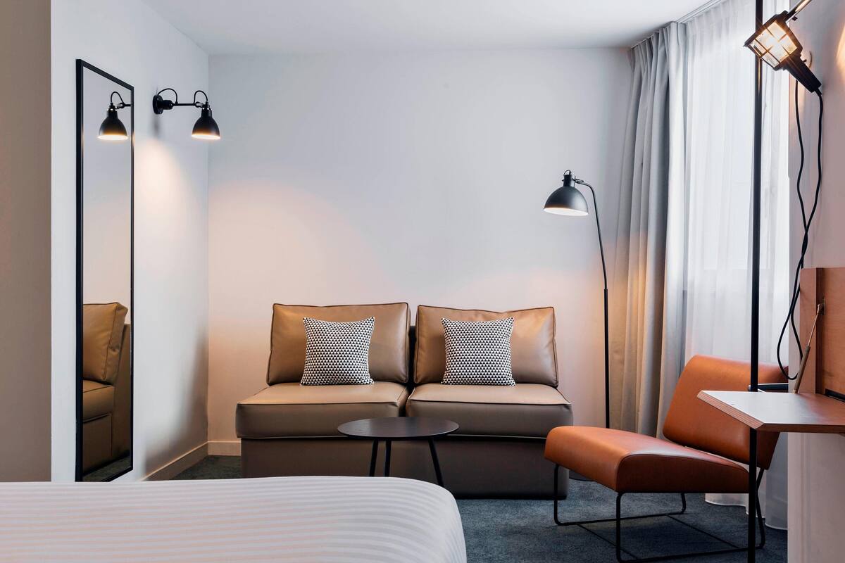 Una habitación de hotel con cama, sofá con dos almohadones, una mesita, una silla, escritorio, lámpara y espejo.