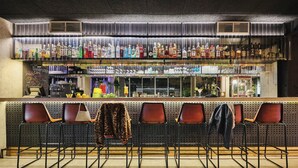 A bar area with a long counter, multiple shelves of bottles, and several chairs with jackets on them. - Moxy Paris Bastille France (Paris)