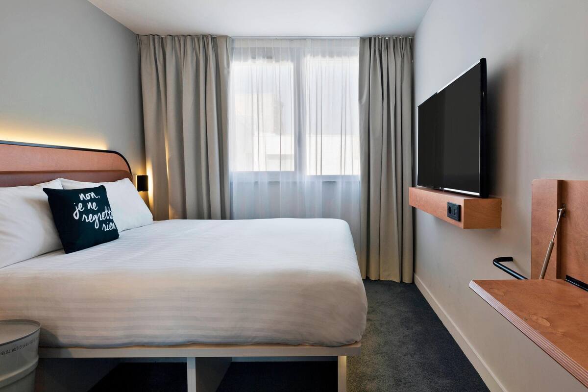 Una habitación de hotel con cama, televisión montada en la pared y ventana con cortinas.
