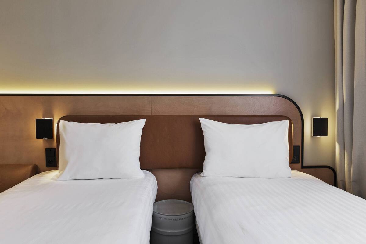 Habitación de hotel con dos camas, cabecero de madera, lámparas de noche y un cubo de basura.