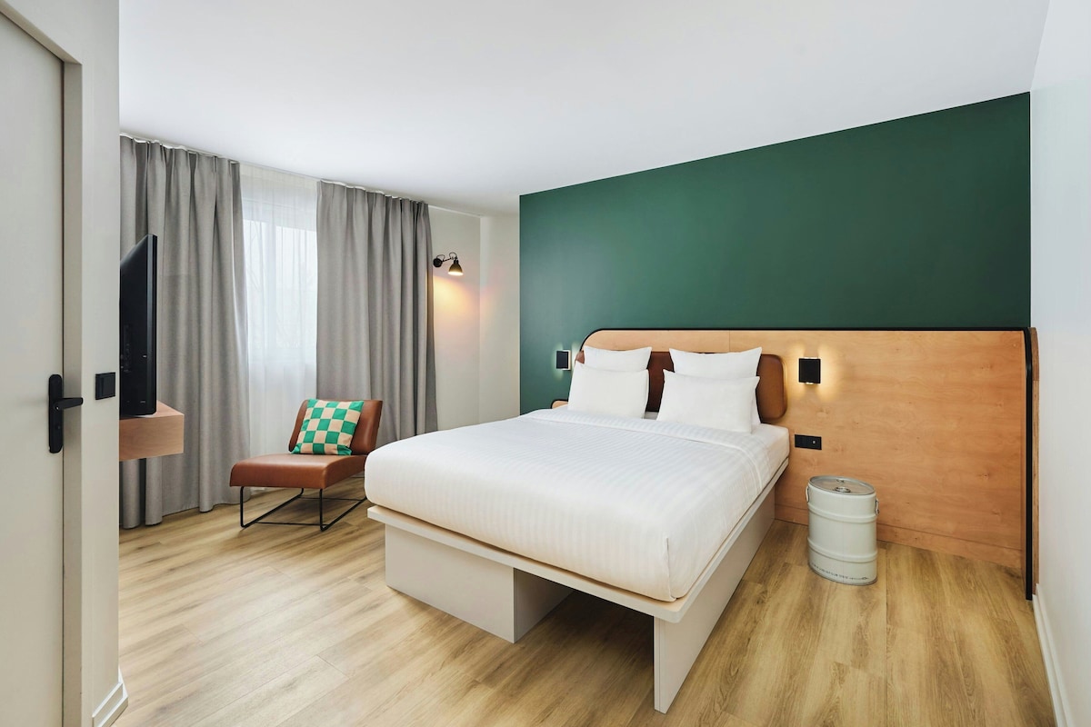 A hotel room with a bed, a chair, a television, and a trash can.