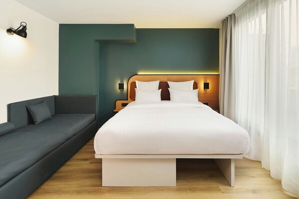 A modern hotel room with a large bed, a dark sofa, and a wall-mounted light fixture.