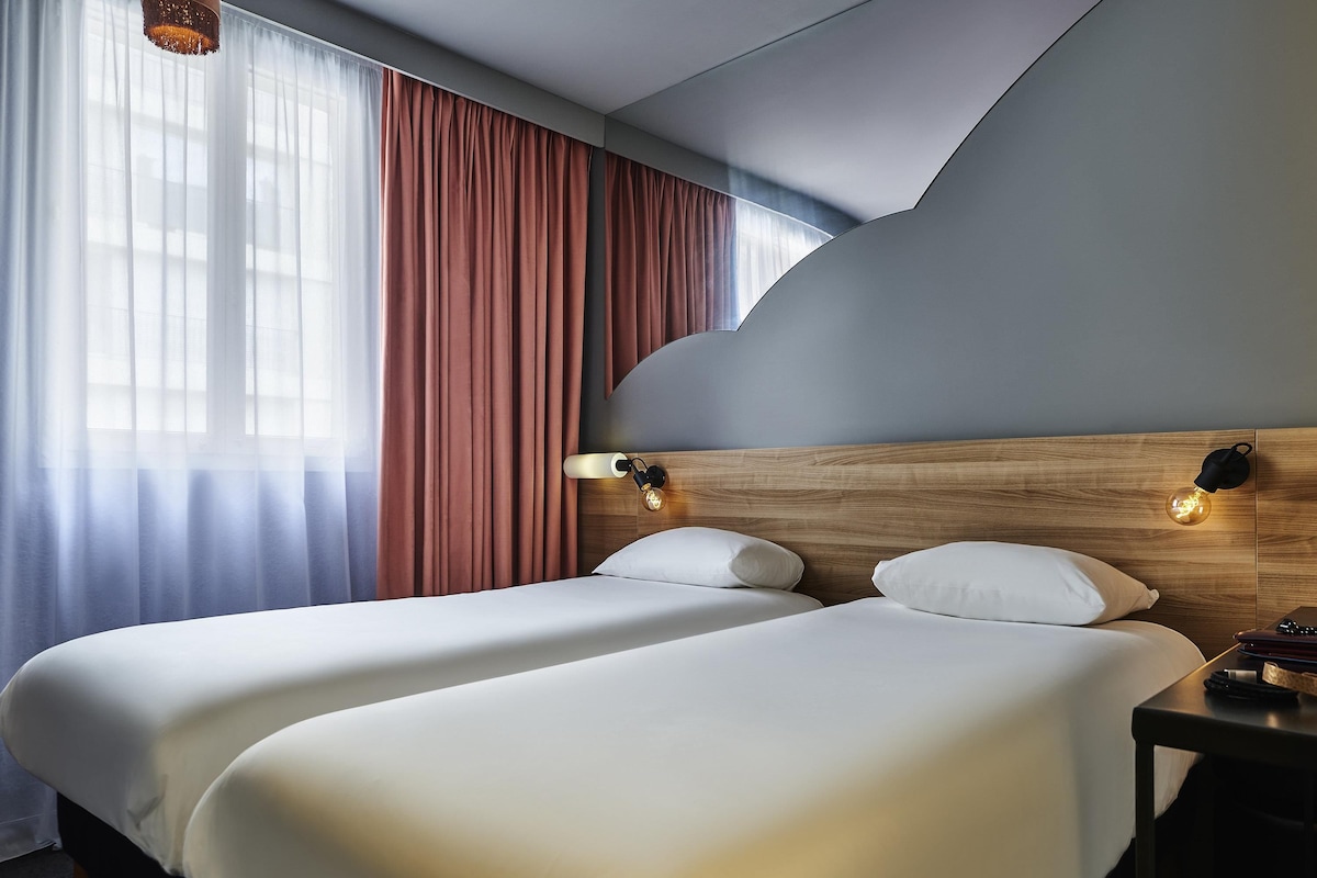 Een hotelkamer met twee bedden, een houten hoofdbord, een nachtkastje en een raam met gordijnen.